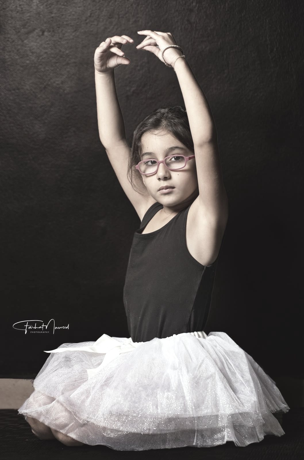 A young ballerina in a black leotard and white tutu, arms raised overhead, studio portrait.