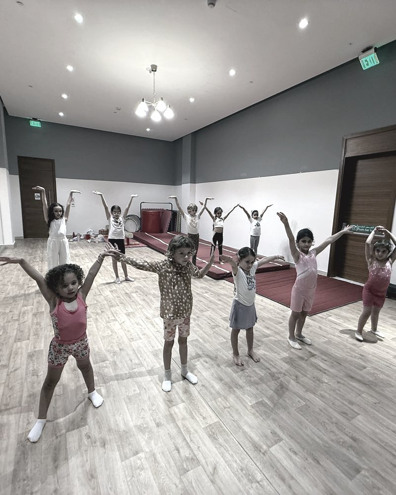 Children stand on a wooden floor in a studio with their arms raised above their heads, red floor mats along the back wall.