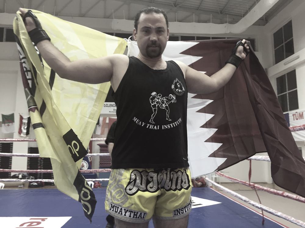 Rami Al Banna holding the Qatari flag open above his shoulders in a sports hall, wearing a Muay Thai Institute vest.