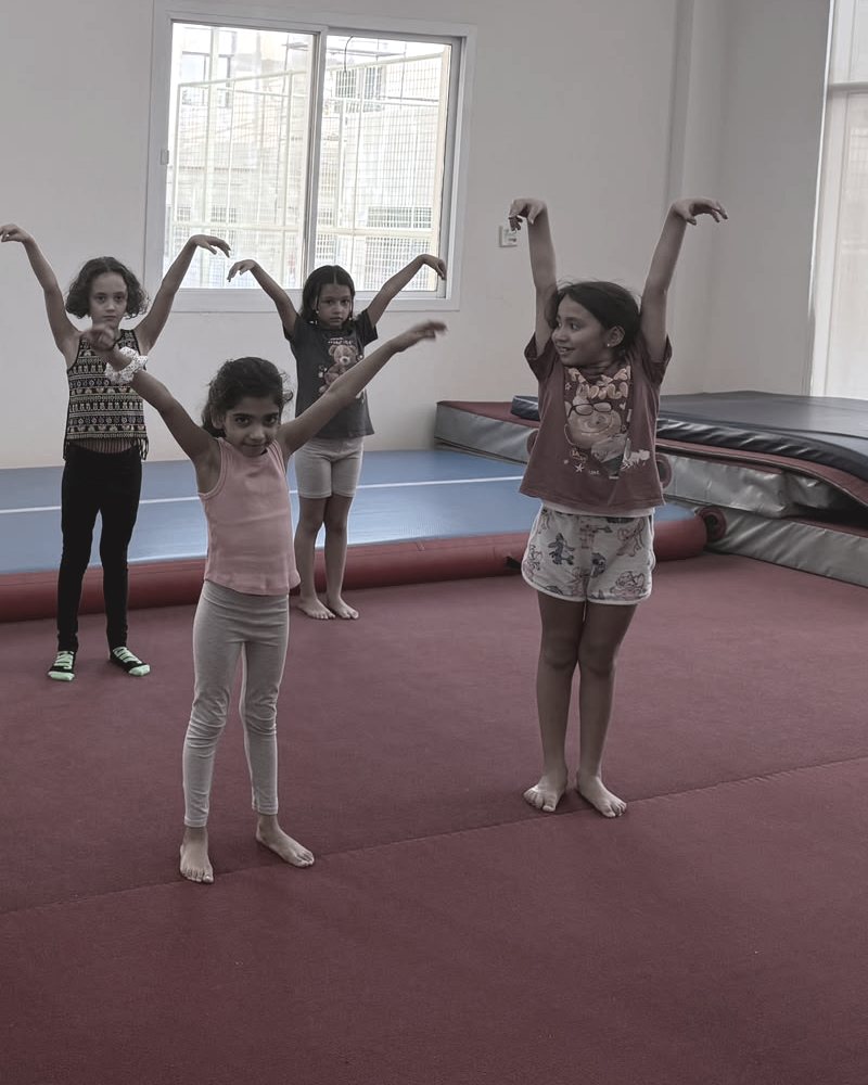 Children stand on red floor matting in a bright room, arms raised above their heads, a blue inflatable mat along the back.