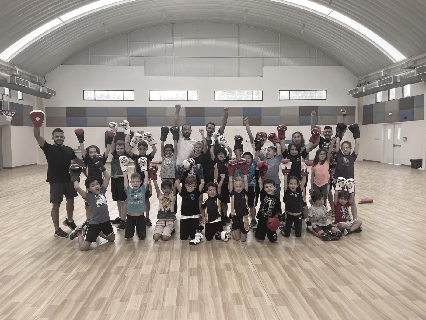 A large mixed class of children and adults training together on a sports hall floor under a high arched roof.