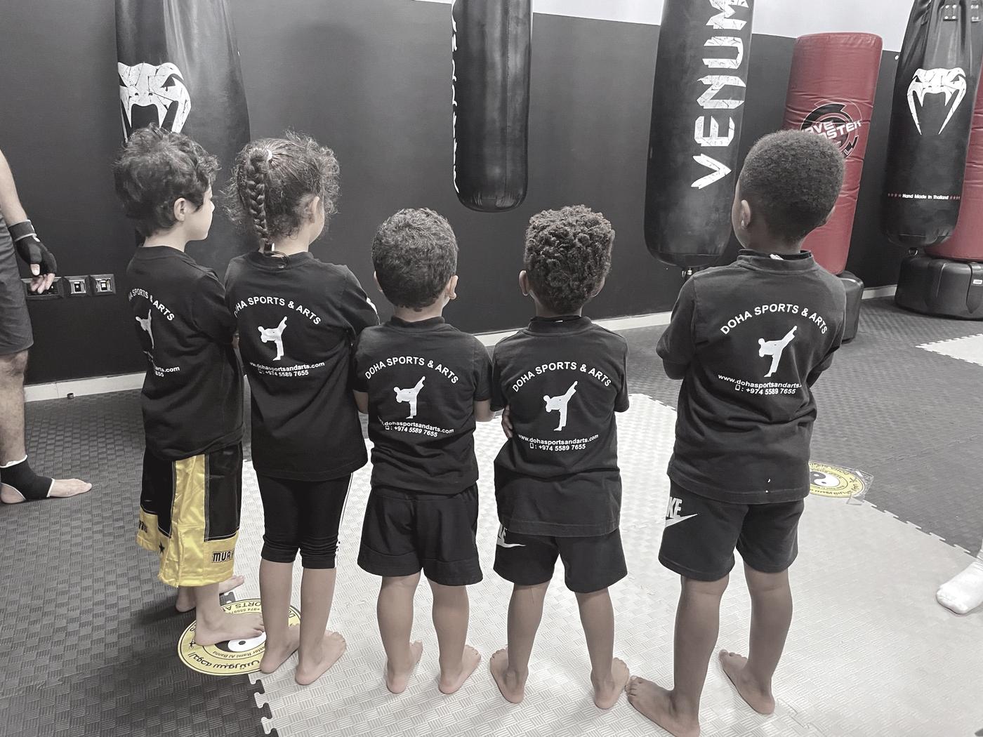 A row of young children photographed from behind in club t-shirts, standing in front of the punching bags.