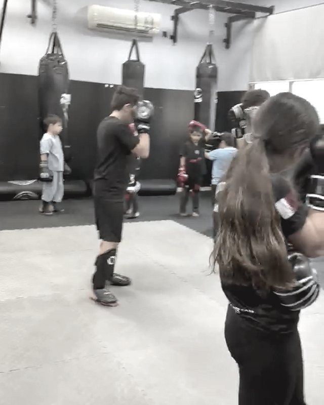 Children in gloves, some in shin guards, work on mats in front of hanging punching bags.