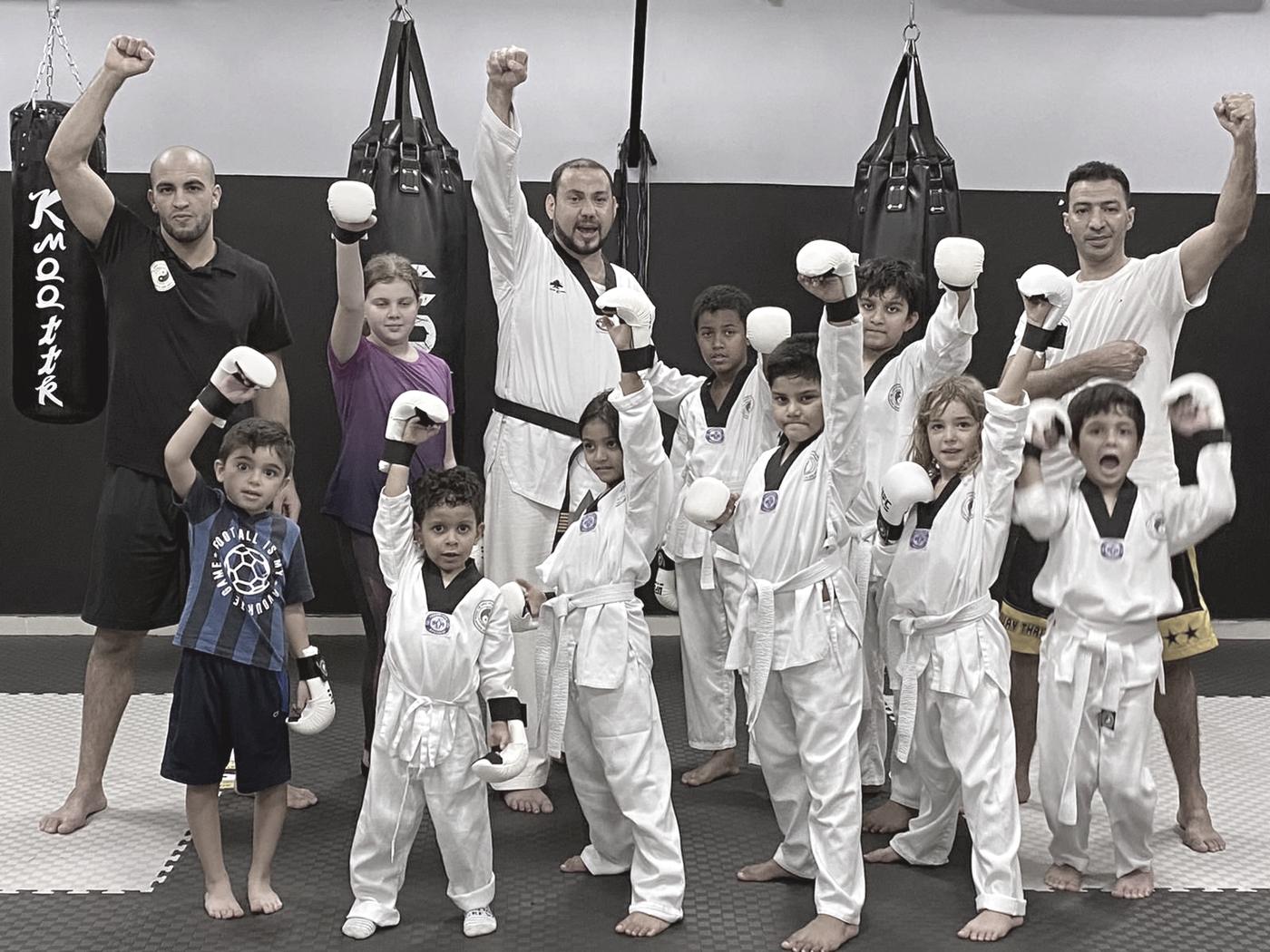 A group of children in white gis with their coaches, arms raised, punching bags hanging behind them.