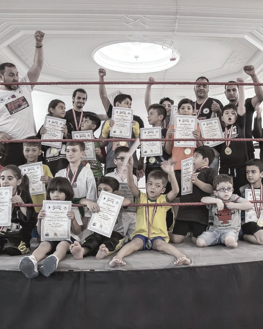 Rows of children and their coaches inside a boxing ring, each child holding a Doha Sports and Arts certificate, fists raised.