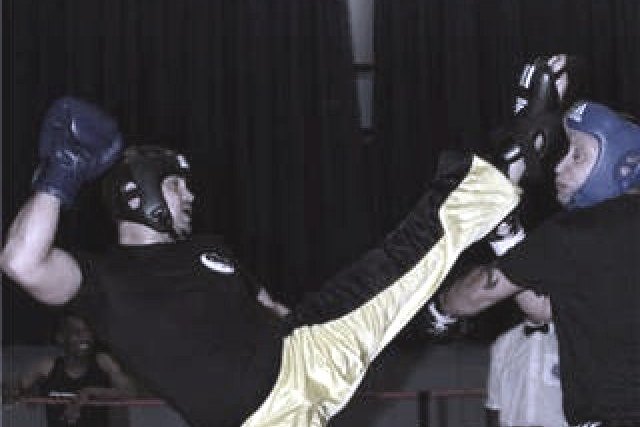 A kickboxer landing a high kick on his opponent's head guard in the ring, both fighters in gloves and headgear, an official standing behind them.