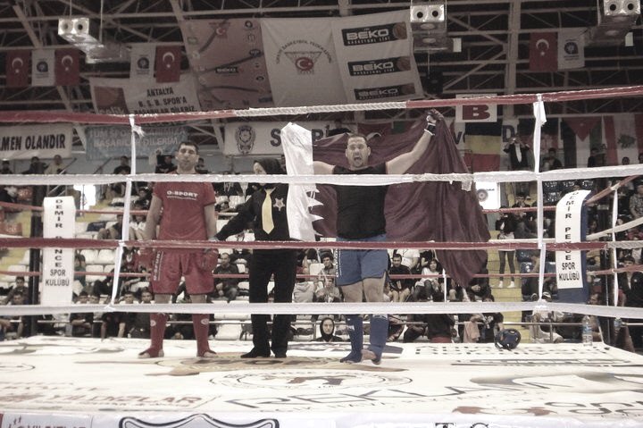 A boxing ring seen from the seats with fighters and officials inside and national flags on the wall behind.
