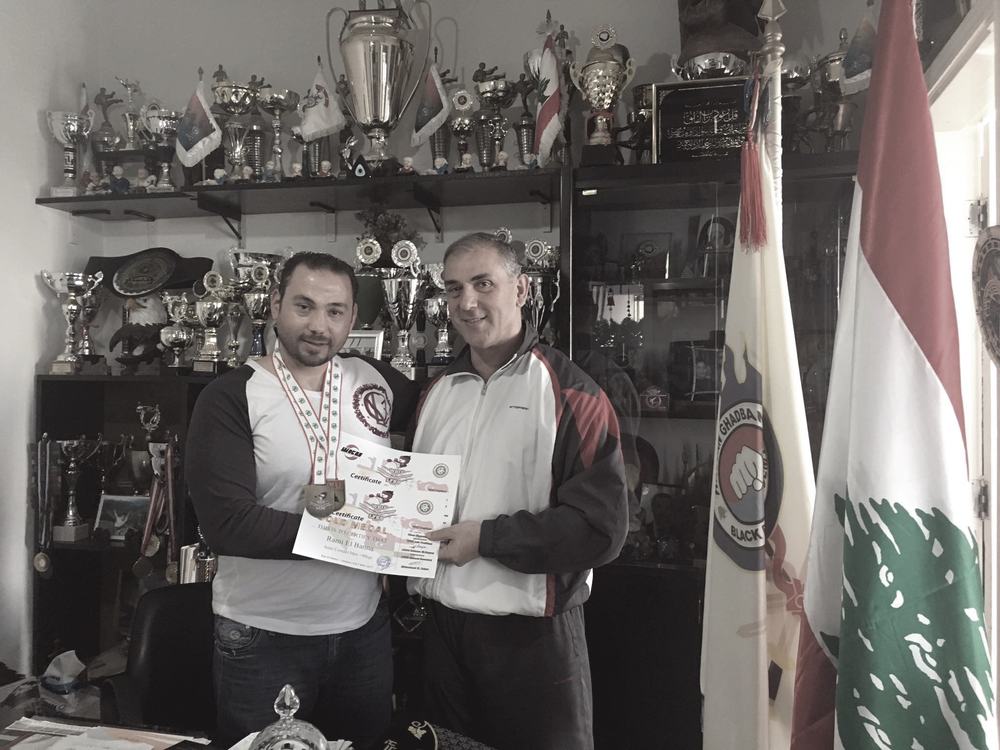 Rami Al Banna receiving a framed award from an older man in a room lined with trophy cabinets, Lebanese flags behind them.