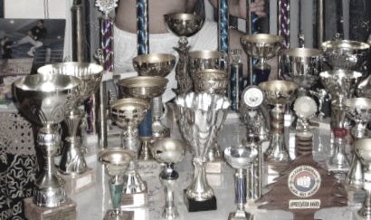 A wall of kickboxing trophies and medals, close together, gold and silver finishes catching the light.
