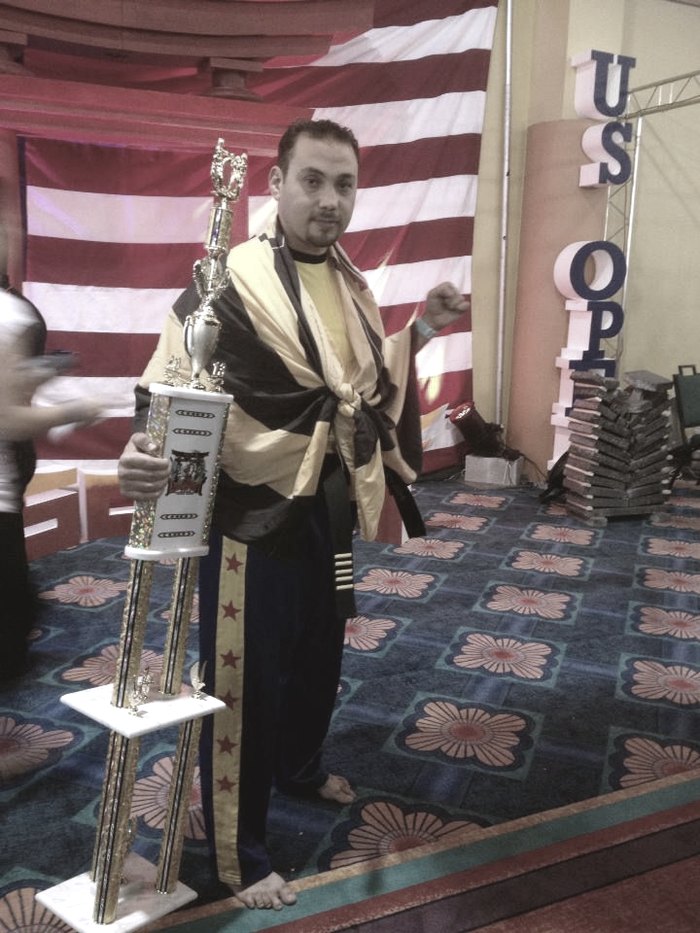 Master Rami Al Banna holding a tall trophy in front of a US Open backdrop, wrapped in a yellow and black sash.