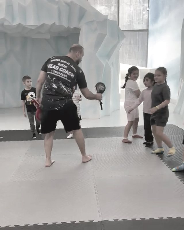 Master Rami Al Banna, in a shirt that reads Head Coach, holds up pads for a child to kick while other children stand behind him, on grey and black mats in front of a blue and white ice-cave backdrop.